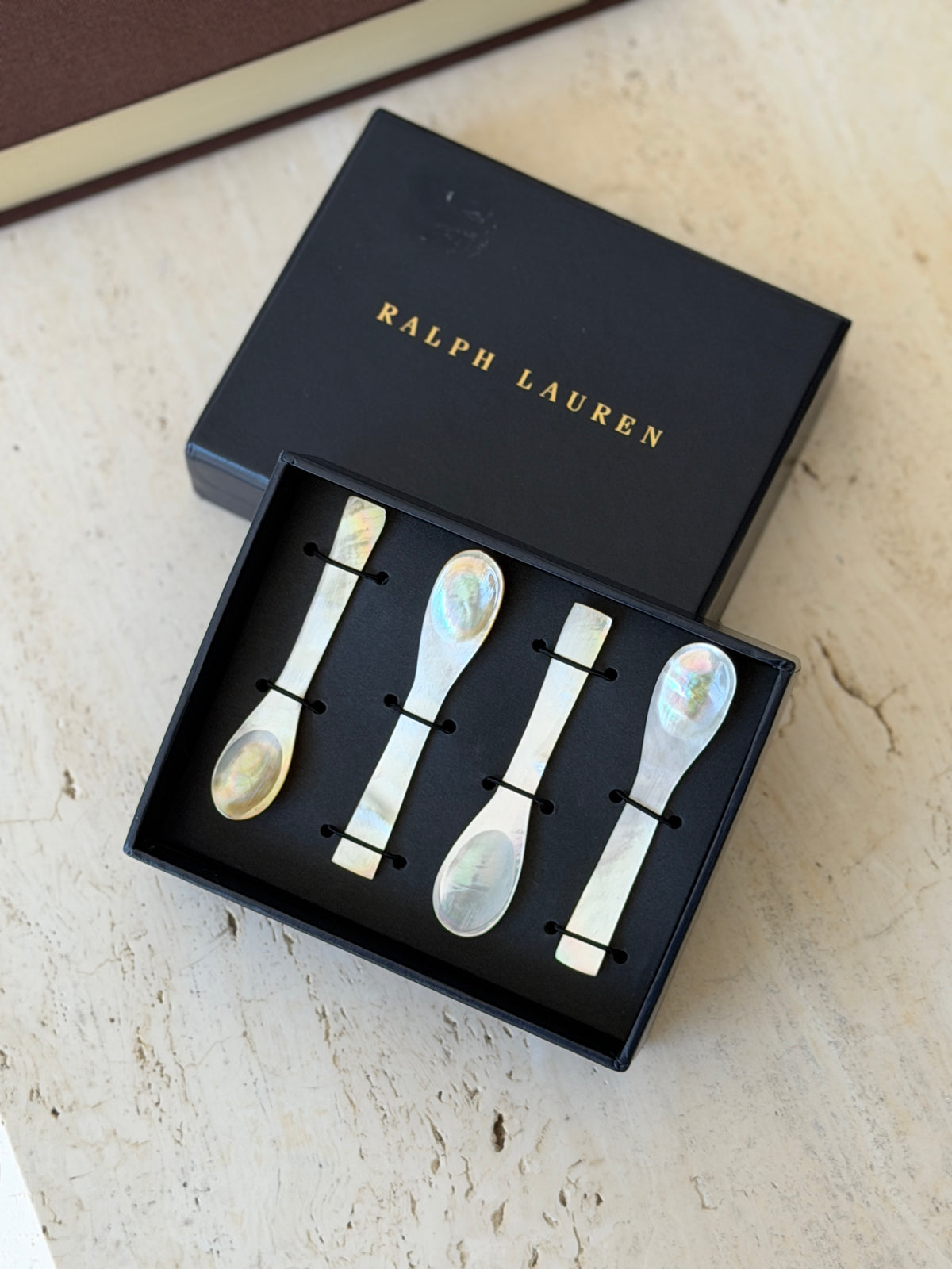 Four mother of pearl caviar spoons secured with black elastic in an open Ralph Lauren box, the gold stamped lid set behind on travertine