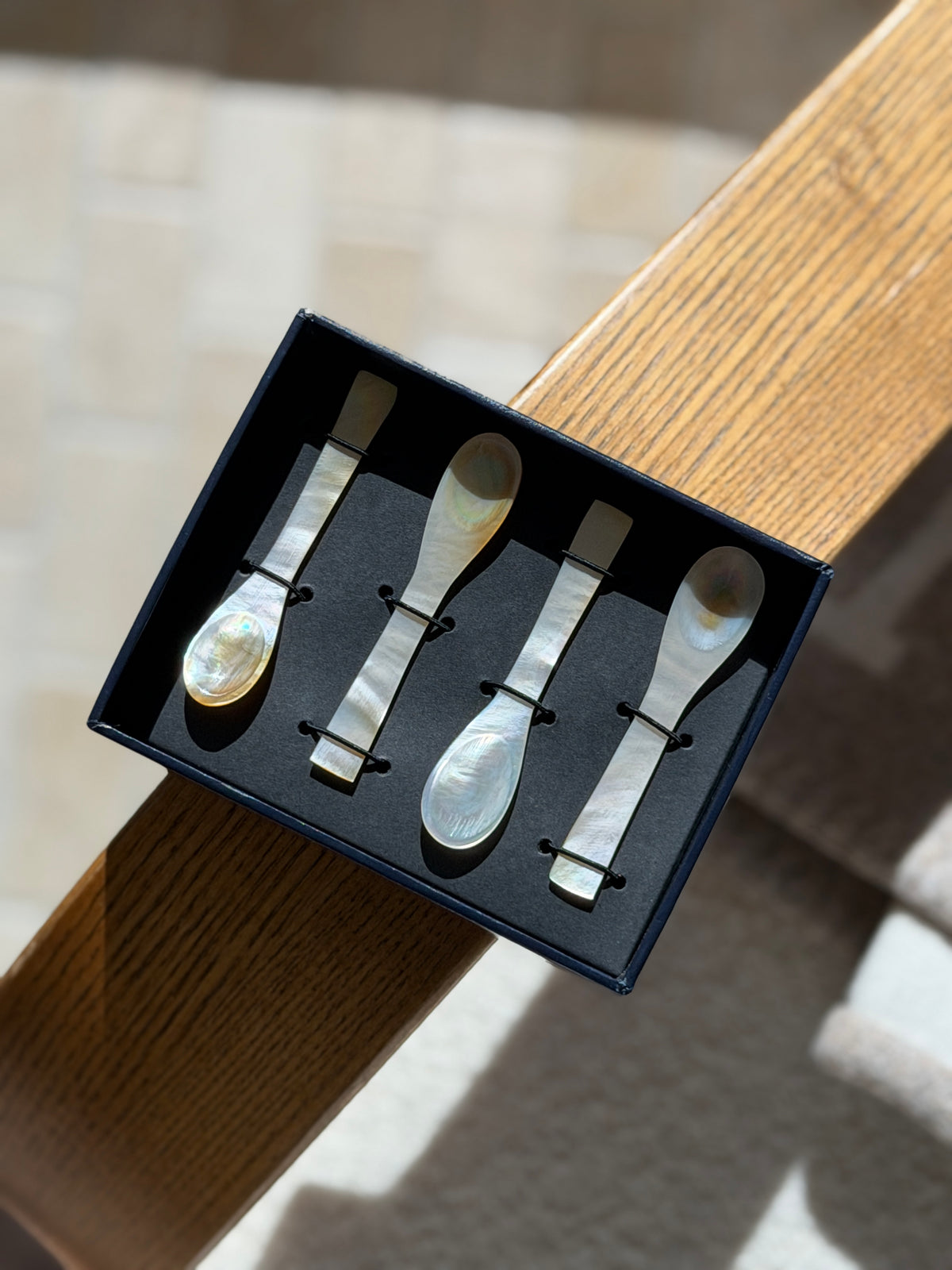 Open box of Ralph Lauren caviar spoons balanced on an oak chair arm in sunlight, the iridescent shell bowls glowing