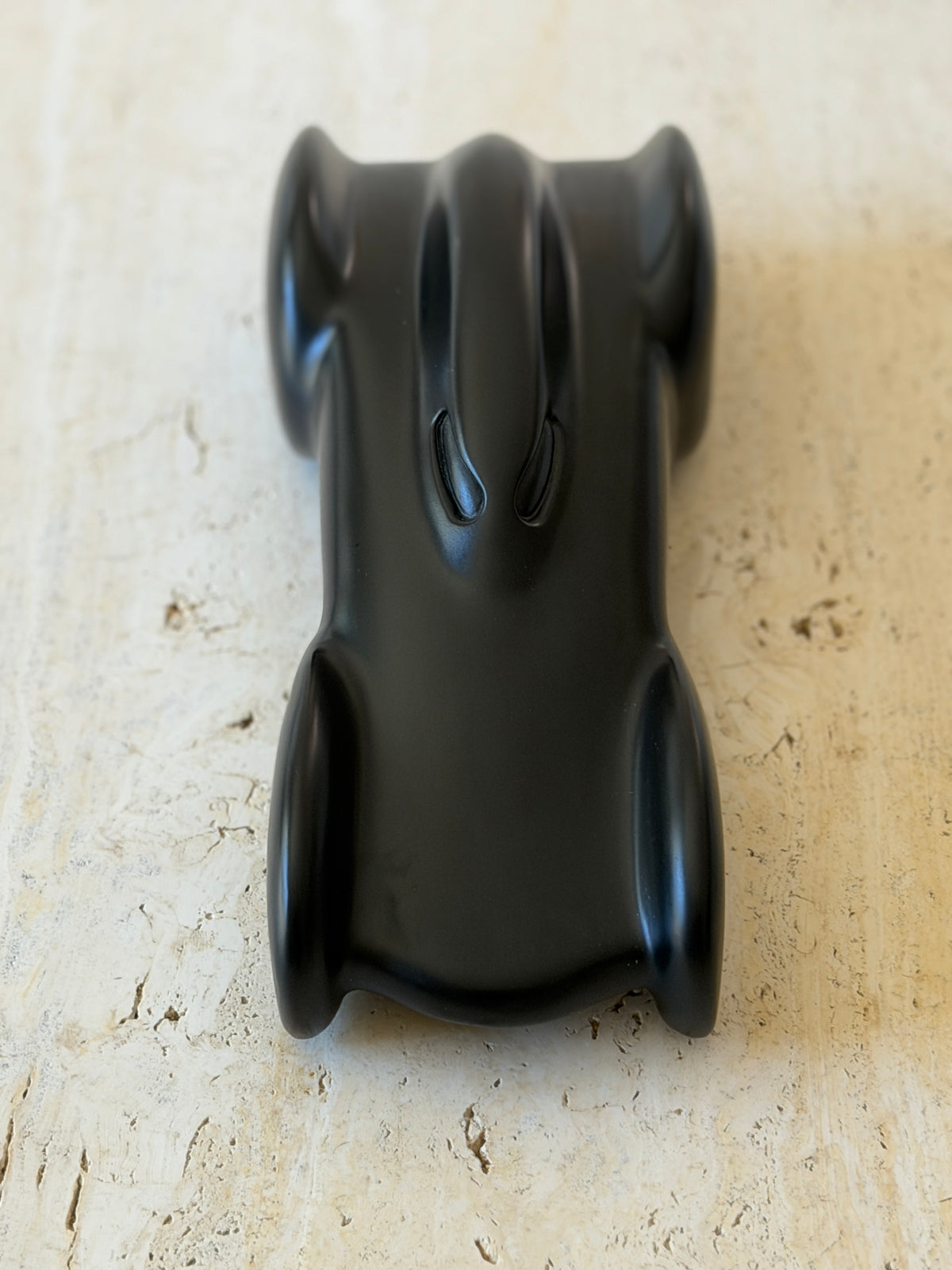 Top down view of the matte black race car sculpture, showing the narrow center body, two slotted cockpit details, and four flared wheel pods