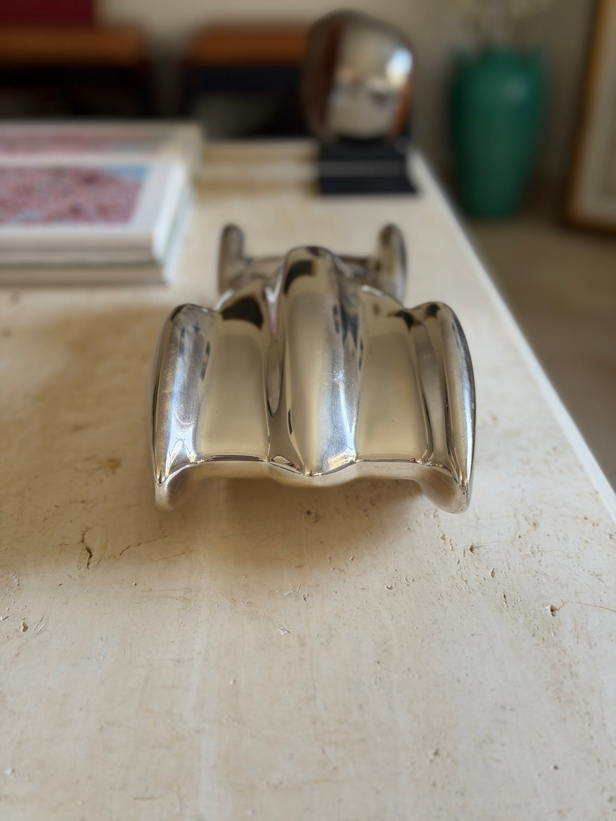 Head on view of the Clermont race car along the travertine table, the sculpted fenders and center hood in mirror finish with stacked books beyond