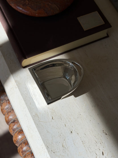 Ralph Lauren stirrup bowl on travertine beneath a brown book and red stone ashtray, the polished silver reflecting the window light