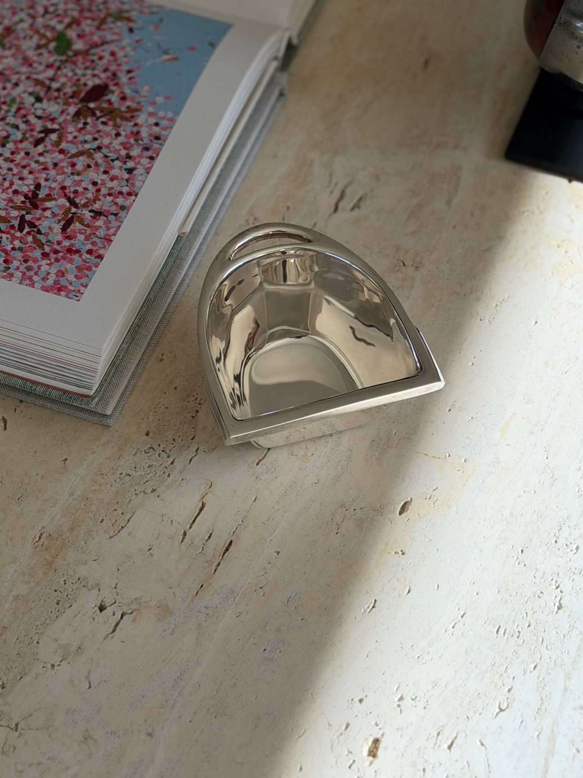 Stirrup shaped silver nut bowl beside an open book of floral paintings, the curved arch and strap slot facing the camera