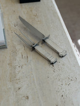 Venturi carving fork and knife angled across a pale travertine surface, their curved crossguards, steel blade, and tines seen in profile