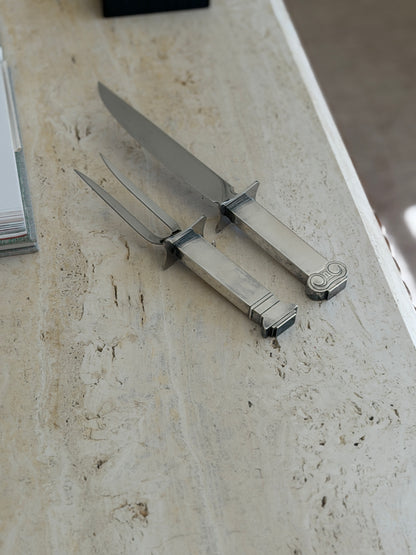 Venturi carving fork and knife angled across a pale travertine surface, their curved crossguards, steel blade, and tines seen in profile