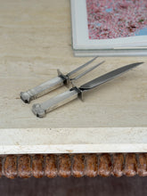 Low angle view of the Robert Venturi carving set, its architectural handles and polished crossguards resting near the edge of a travertine tabletop
