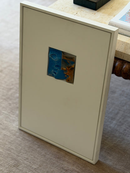 Full view of the tall white shadowbox frame leaning against a travertine console with a carved wood base, the small bronze relief set in its upper window