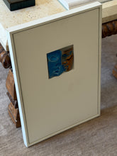 Angled view of the framed bronze relief against a travertine console, showing the recessed window and the deep white frame profile