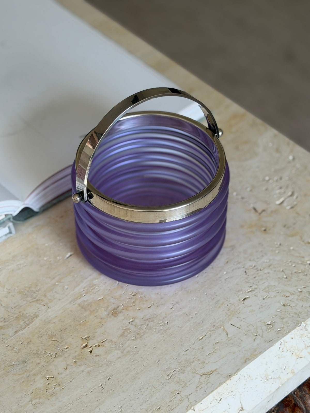 Overhead angled view of the purple ribbed Onde ice bucket with its polished silver swing handle raised, resting on a stone surface beside an open book