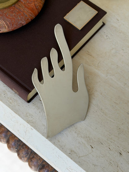 Flat silver metal hand sculpture with elongated wavy fingers, leaning against a brown book on travertine