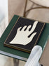 Silver hand sculpture on a black and gold bordered book atop an Audemars Piguet volume, a frosted glass object in the foreground