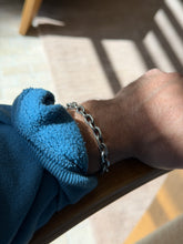 Oval link bracelet on a wrist resting on a wooden chair arm, striped sunlight falling across the floor behind