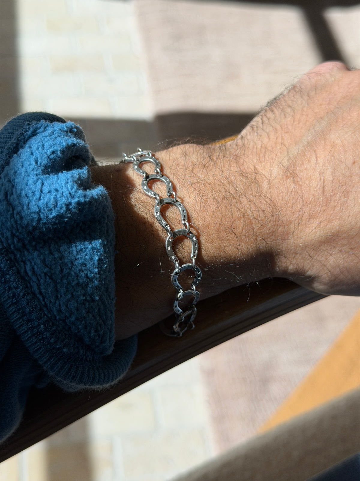 Horseshoe link bracelet on a wrist resting on a wooden chair arm, strong sunlight and shadow across the silver links