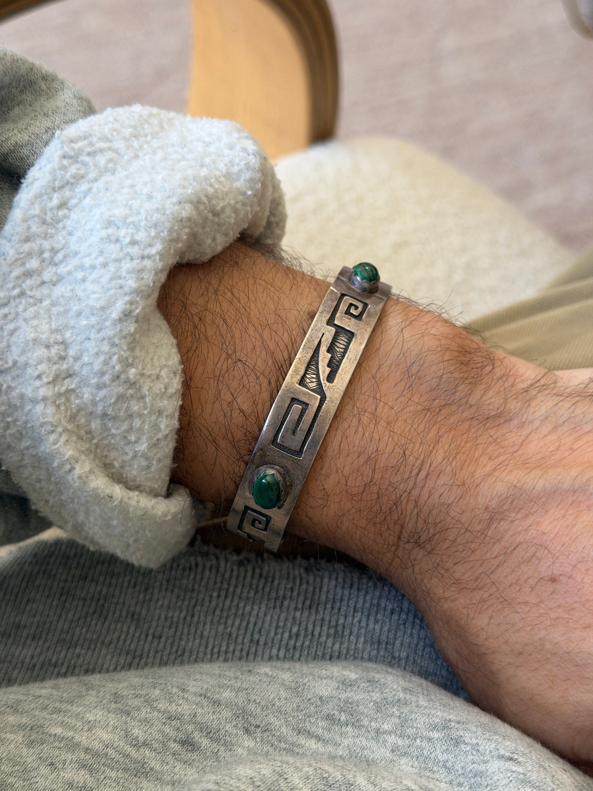Malachite and sterling silver cuff on the wrist beside a bentwood chair, showing the scalloped bezels and hatched texture within the recessed design