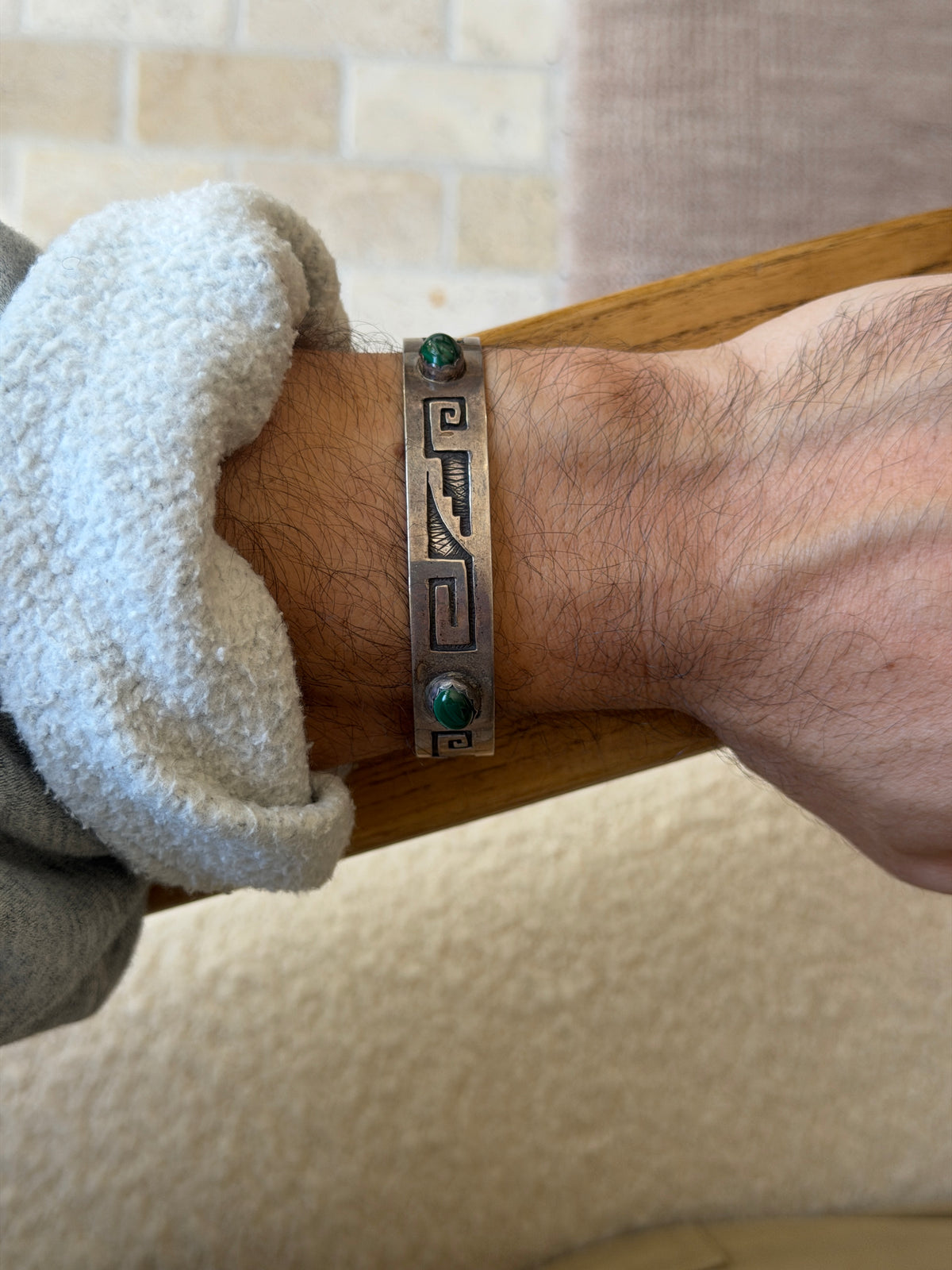 Straight on view of the sterling cuff worn against a wooden chair arm, its key pattern and banded green malachite stones centered on the wrist