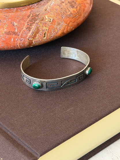 Open sterling silver cuff resting on a brown clothbound book beside a red stone dish, revealing its plain interior and both malachite cabochons