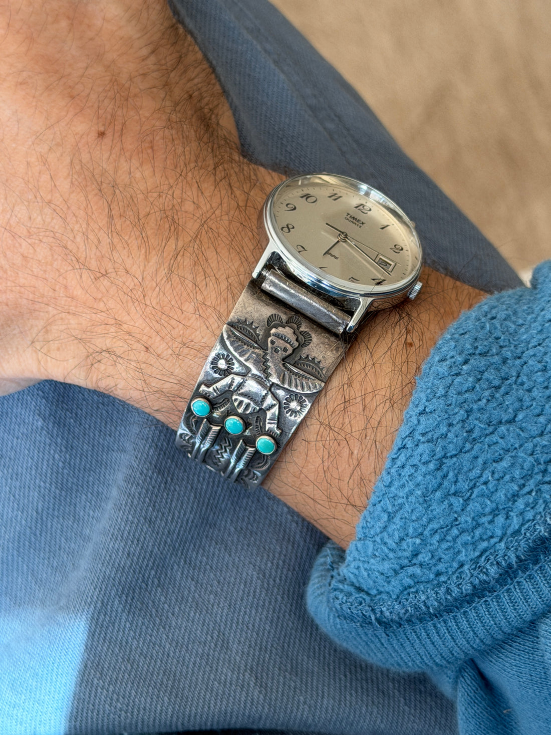 Angled view of the Native American watch cuff, showing the stamped winged dancer figure above a row of three bezel set turquoise stones