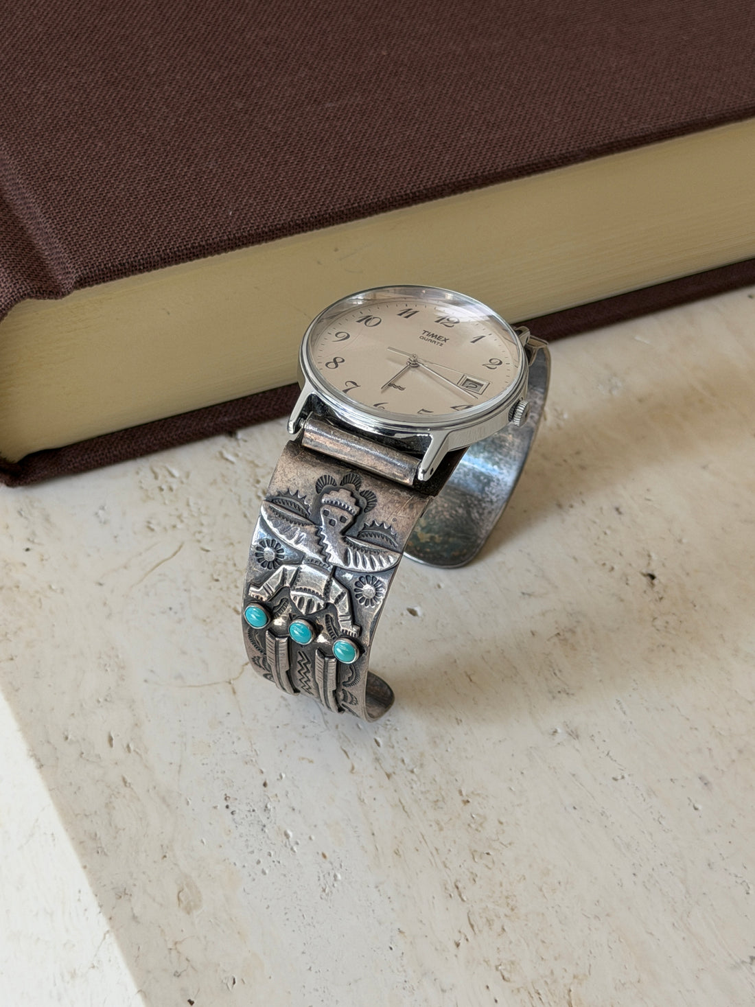 Sterling silver watch cuff standing on travertine beside a brown clothbound book, the winged figure and turquoise trio facing forward