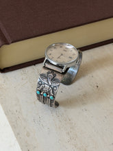 Sterling silver watch cuff standing on travertine beside a brown clothbound book, the winged figure and turquoise trio facing forward
