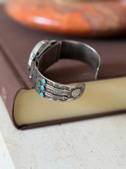 Side view of the watch cuff resting on a brown book, the arrows, turquoise stones, and sun stamp running along the band