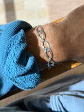 Oval figure eight bracelet on a wrist resting on a wooden chair arm, sun catching the engraved silver