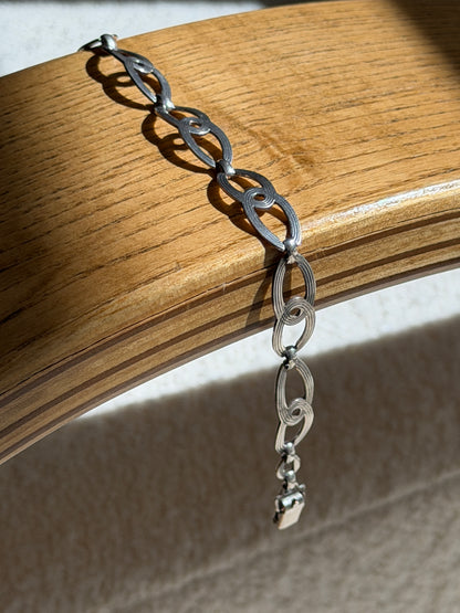 Sterling silver figure eight link bracelet draped over a bentwood oak chair arm, ending in a box clasp