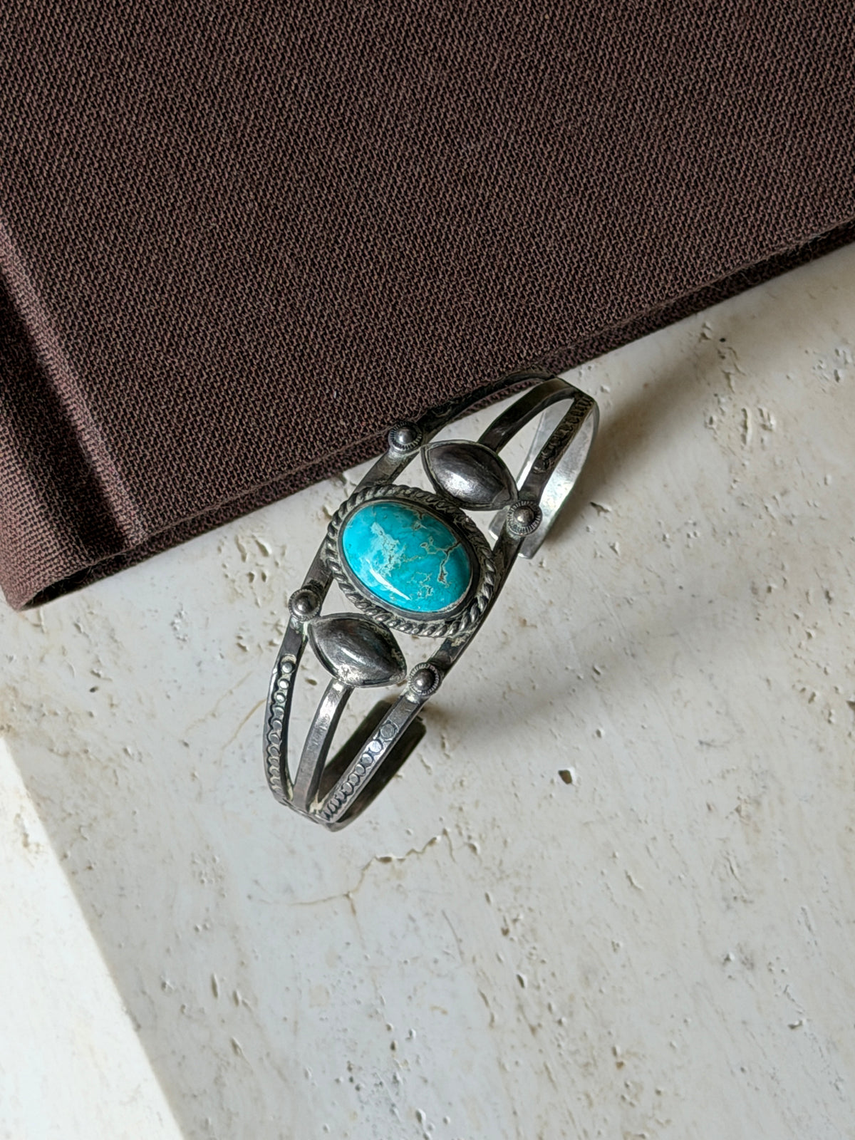 Overhead view of the triple band cuff on travertine beside a brown book, the veined turquoise centered between two domed leaves