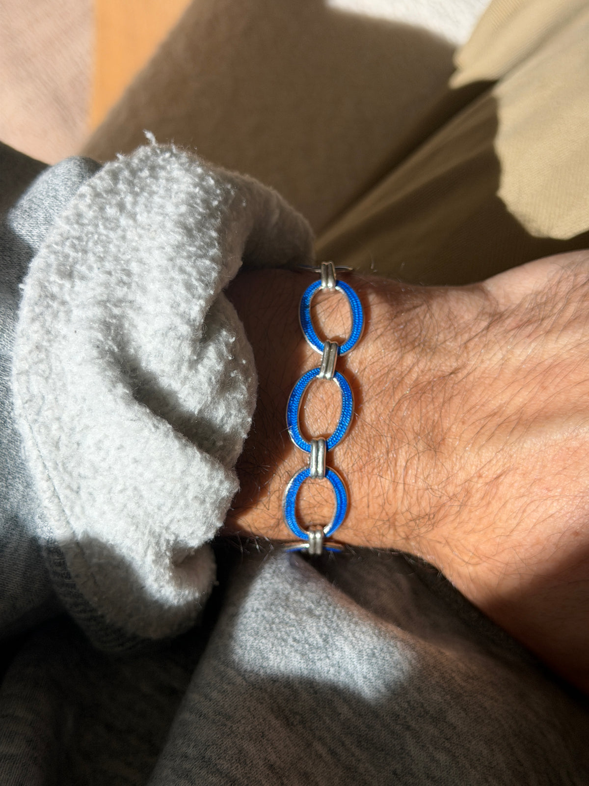 Sterling silver bracelet of oval links in textured blue enamel joined by ribbed silver connectors, worn on the wrist in warm sunlight beside a gray fleece cuff