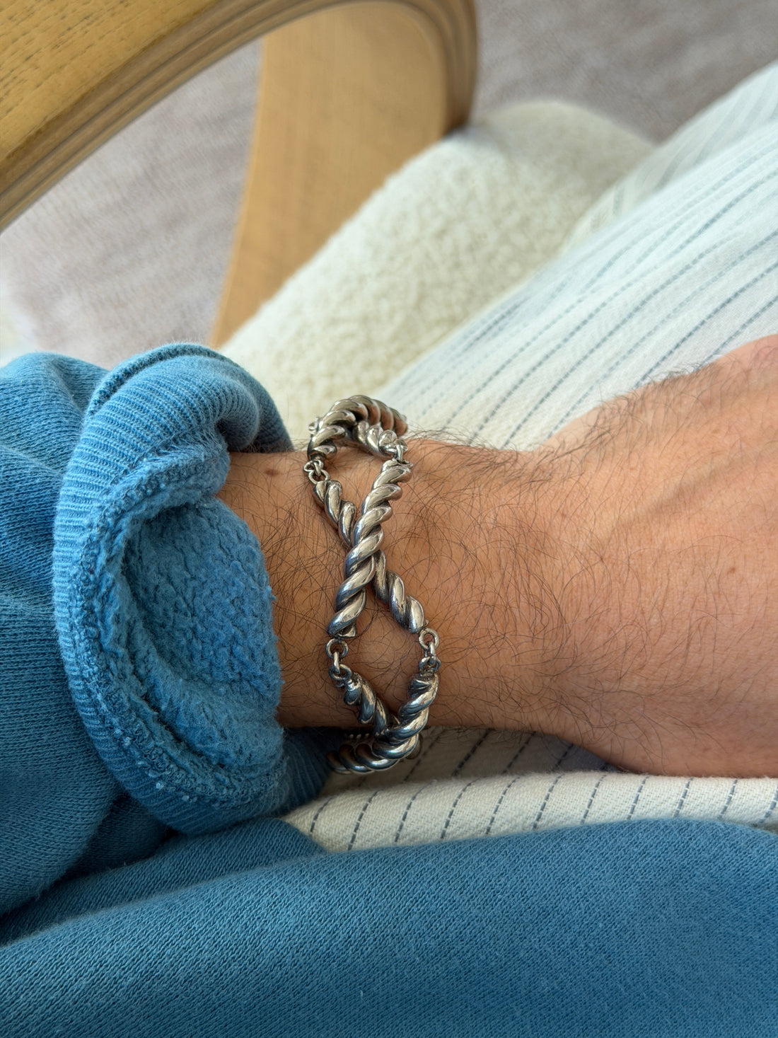 Silver cordage gated link bracelet worn with a rolled blue sleeve, the wrist resting on a striped cushion beside a bentwood chair frame