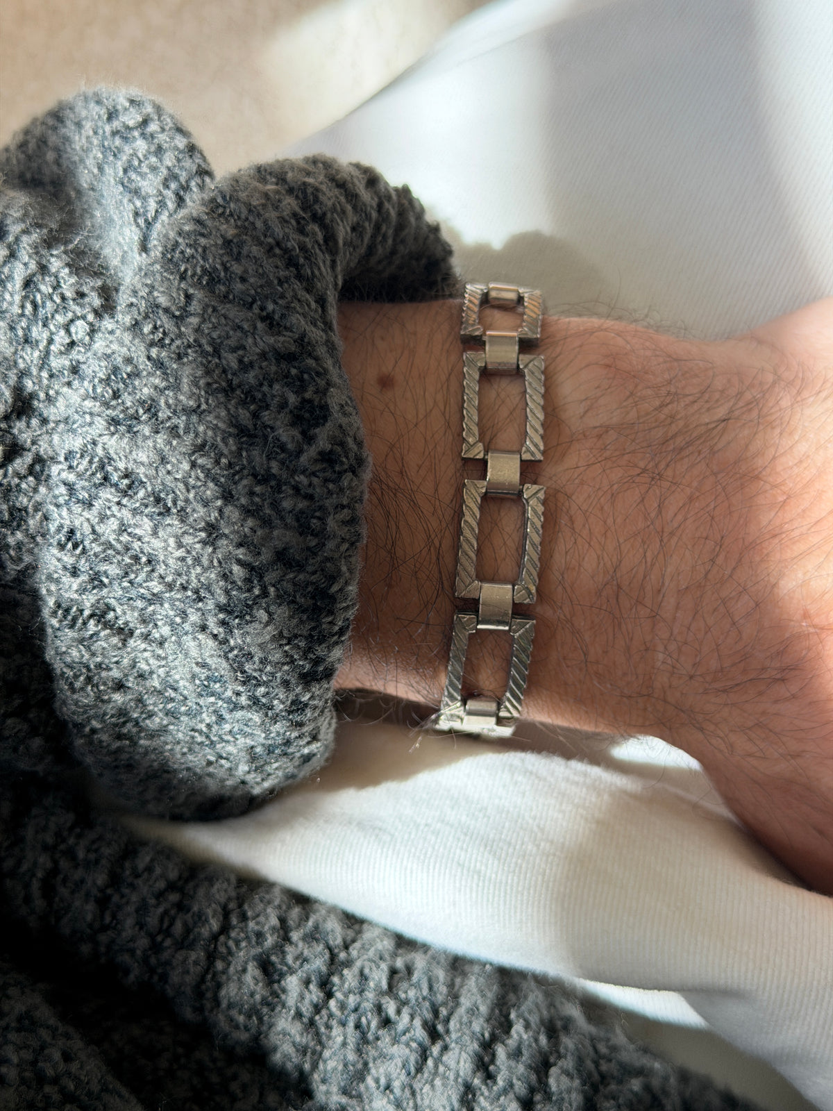 Rectangular panel link bracelet on the wrist against a gray knit cuff, the diagonal twisted rope engraving catching soft light
