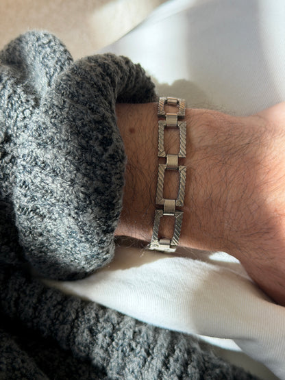 Rectangular panel link bracelet on the wrist against a gray knit cuff, the diagonal twisted rope engraving catching soft light