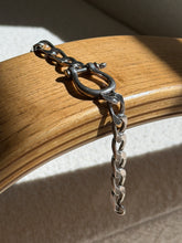 Sterling horseshoe shackle bracelet draped over a bentwood oak chair arm, the curb chain hanging free in raking sunlight