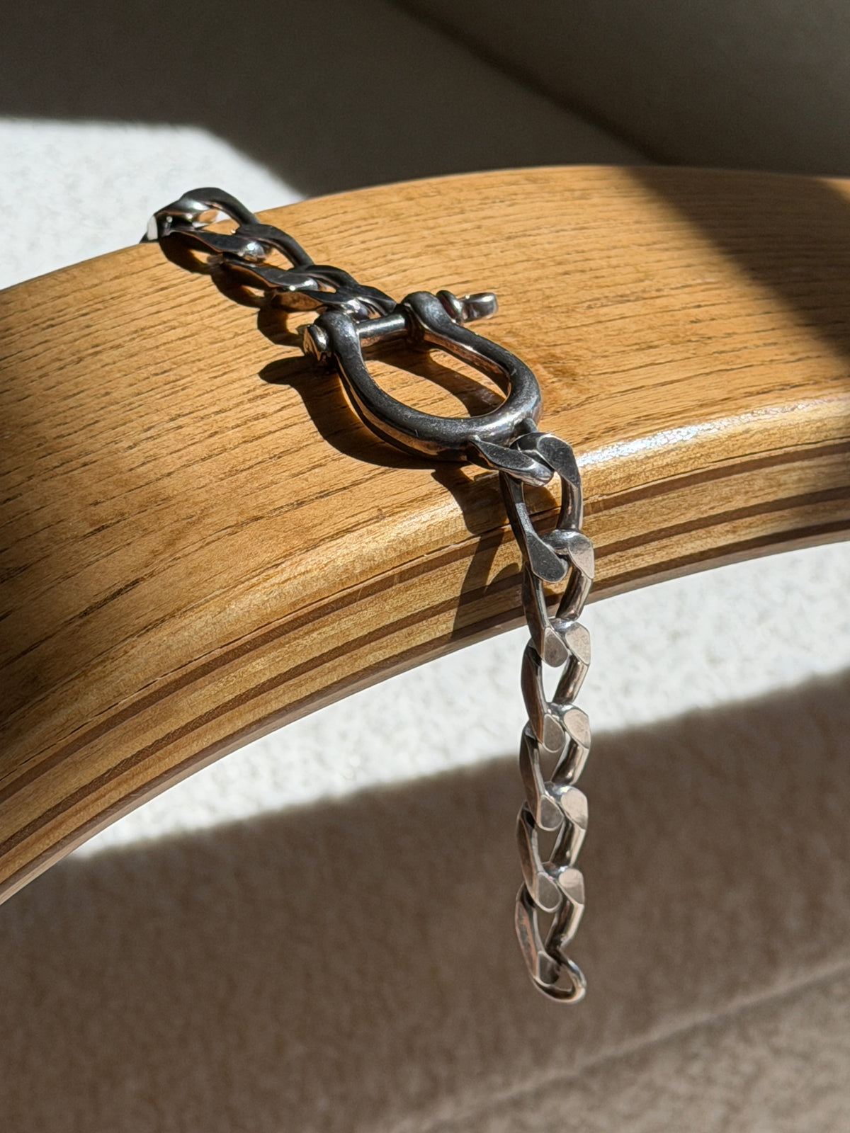 Sterling horseshoe shackle bracelet draped over a bentwood oak chair arm, the curb chain hanging free in raking sunlight