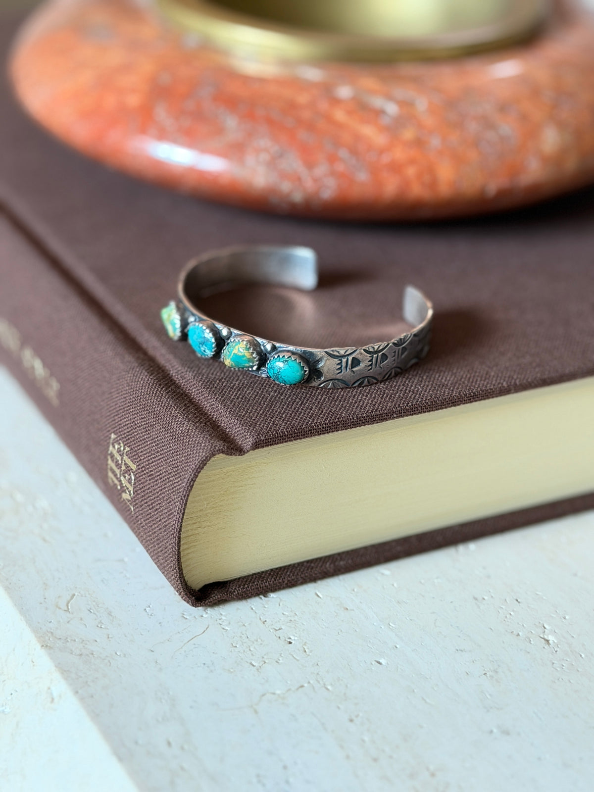 Cuff resting on a brown gold-tooled book, with a blurred rust-colored ceramic dish behind it