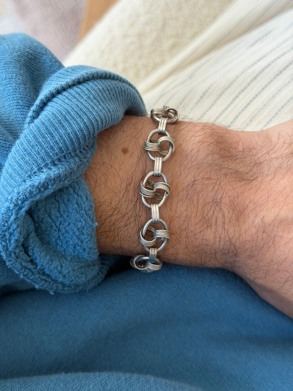 Bracelet worn on the wrist near a wooden chair frame, cream top and blue sweatshirt cuff visible