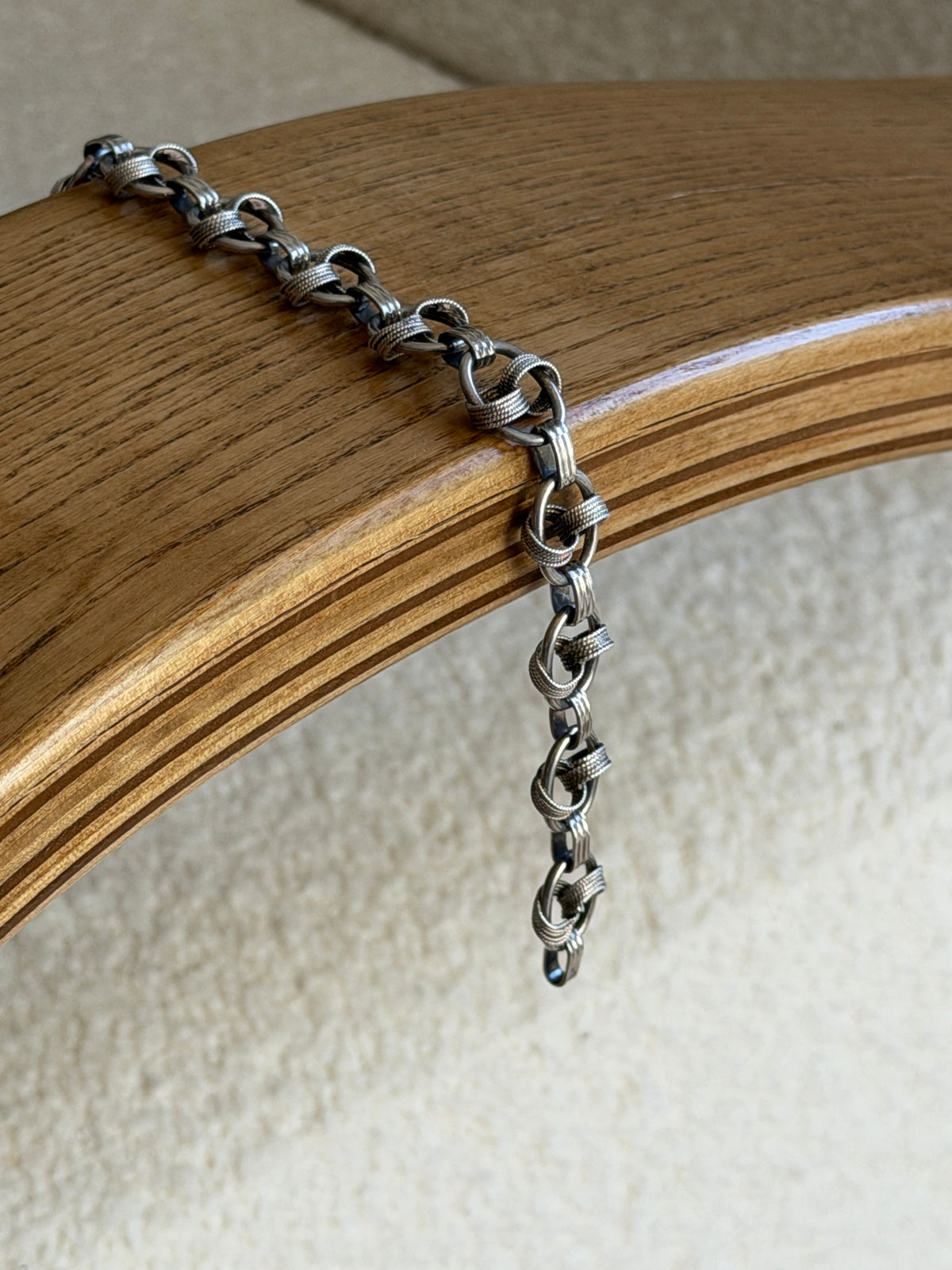 Bracelet draped over the curved edge of a wooden chair arm, showing its fluid articulation