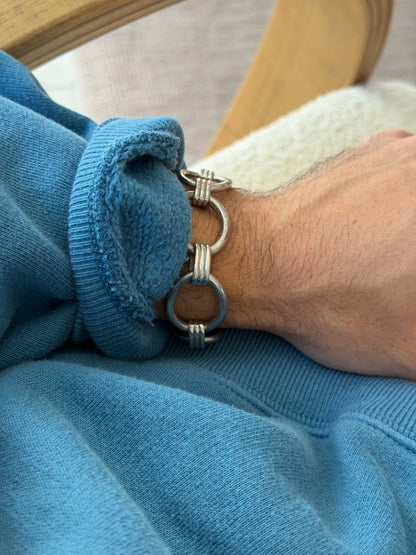 Bracelet worn on the wrist near a wooden chair frame and cream cushion, showing the open oval links clearly