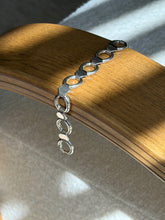Sterling round and hexagon link bracelet draped over a bentwood oak chair arm in striped sunlight, the clasp end hanging below